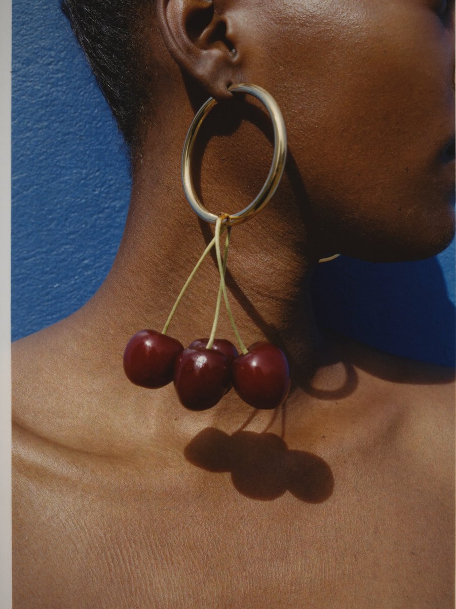 Une créole dorée à laquelle pendent trois cerises, ombre portée nette sur le cou, mur cobalt