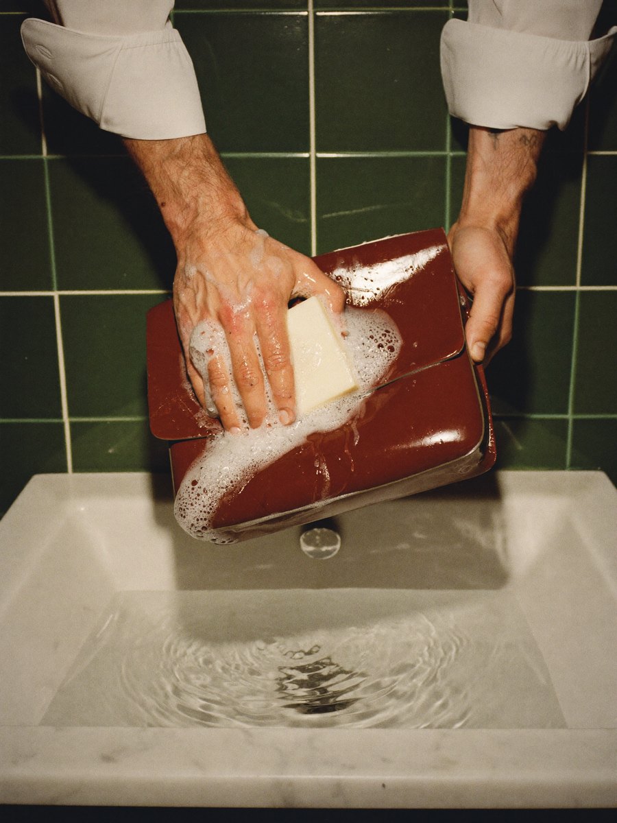 Deux mains d'homme savonnent un sac en cuir verni bordeaux dans un lavabo de marbre